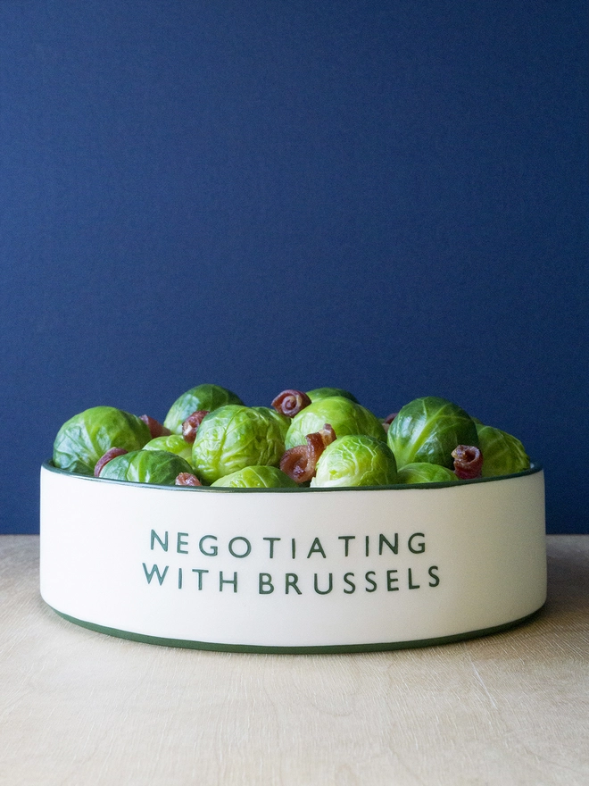 Ceramic porcelain dish with Negotiating with Brussels written on the side. A white Christmas serving dish with green rim containing Brussels sprouts and bacon.