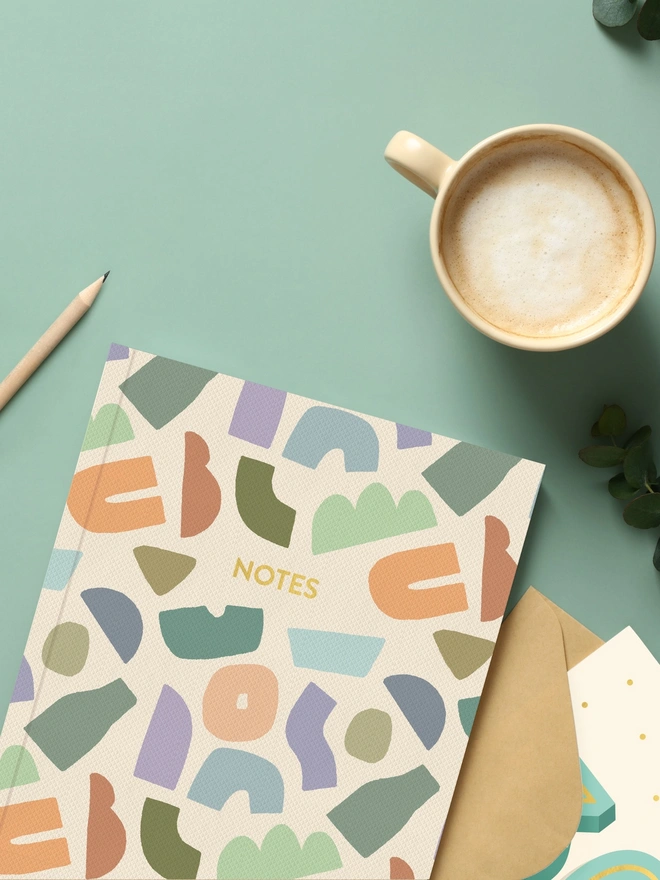 Notebook with abstract shapes on cover sat on a green coloured desk with a mug of coffee and stationery.