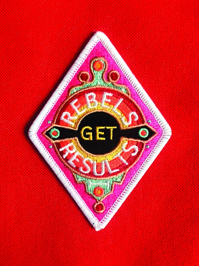 Sitting on a red background is a diamond shape embroidered patch with “Rebels Get Results” at the centre. The design features pink, white, red, yellow and black colours and has a white border.