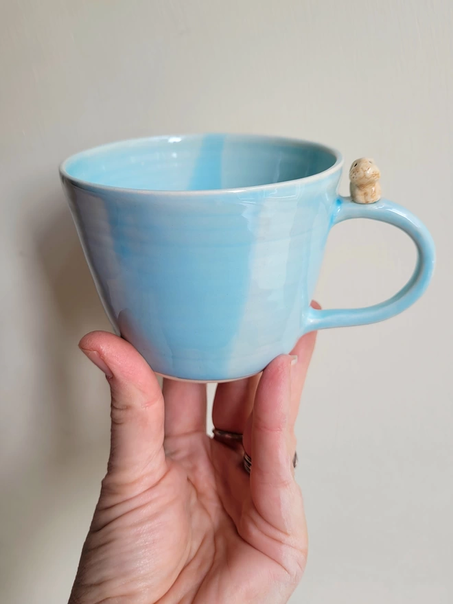 Shiney pale blue cup with beige ceramic bunny rabbit on the handle held in a hand