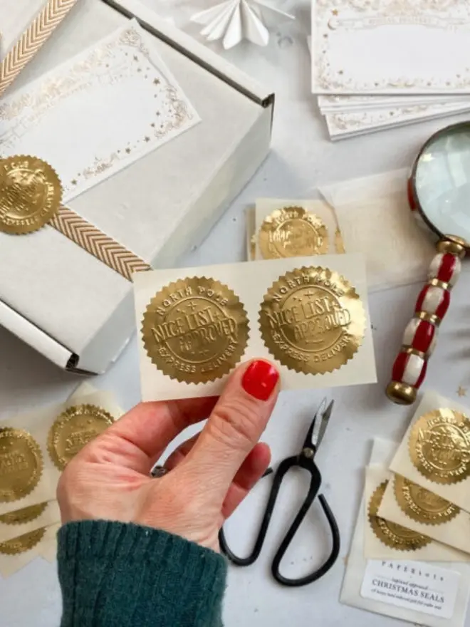 A hand holds a sheet of two gold "Nice List Approved" seals. More gold seal sheets, blank "Special Delivery" labels and a wrapped box are spread around it.