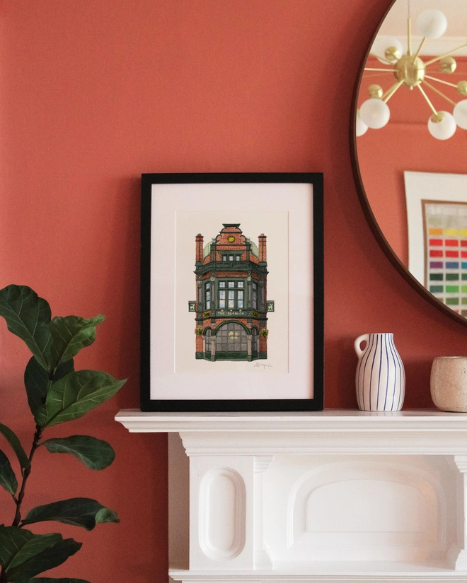 A framed print featuring an illustration of The Roebuck pub displayed on a white mantelpiece against a terracotta-colored wall.