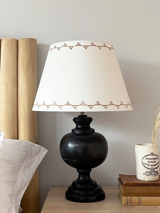 Empire lampshade for table lamps in cream with geometric triangle design, styled on a wooden bedside table with books, black lamp base and white bedding. There is bight orange and white artwork above the bed.