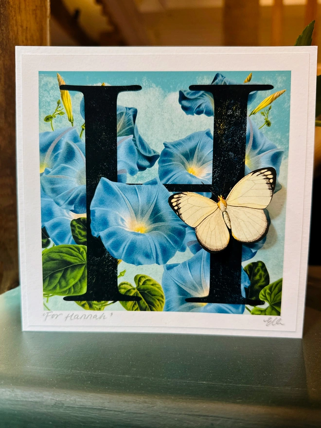 An example of a letter H, birthday, butterflygram, showing the birth flower, morning glory, a blue, trumpet flower and a 3 dimensional white paper cut butterfly on the letter H. Shows card on display on a shelf.