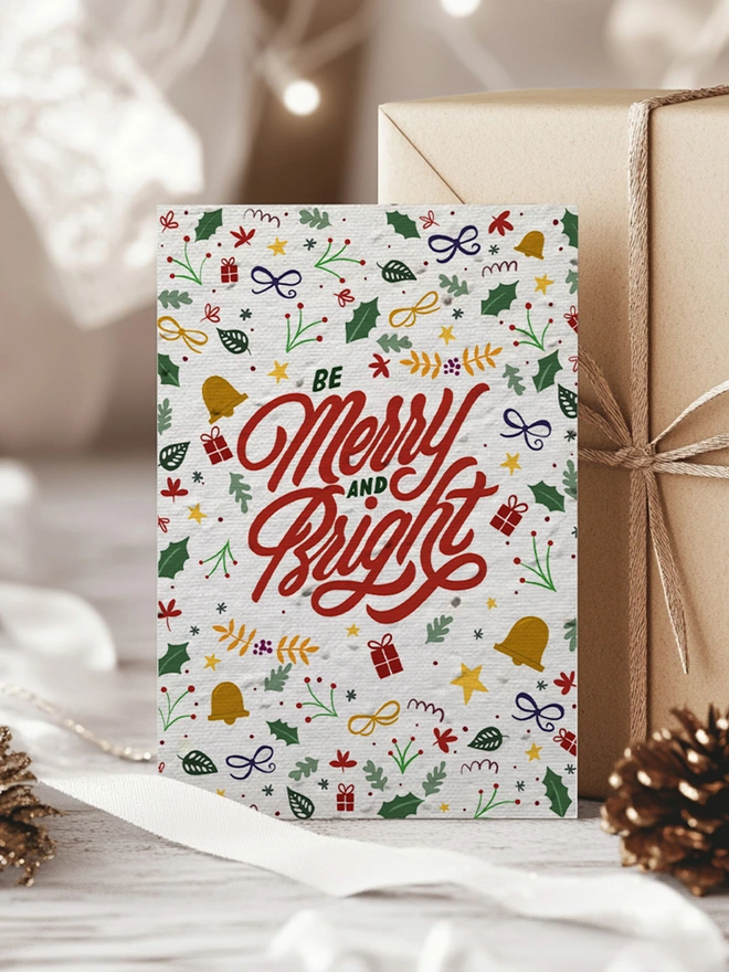 Christmas card with Merry and Bright in the centre with illustrated motifs surrounding the text. The card is on a white washed floor with a present behind the card.