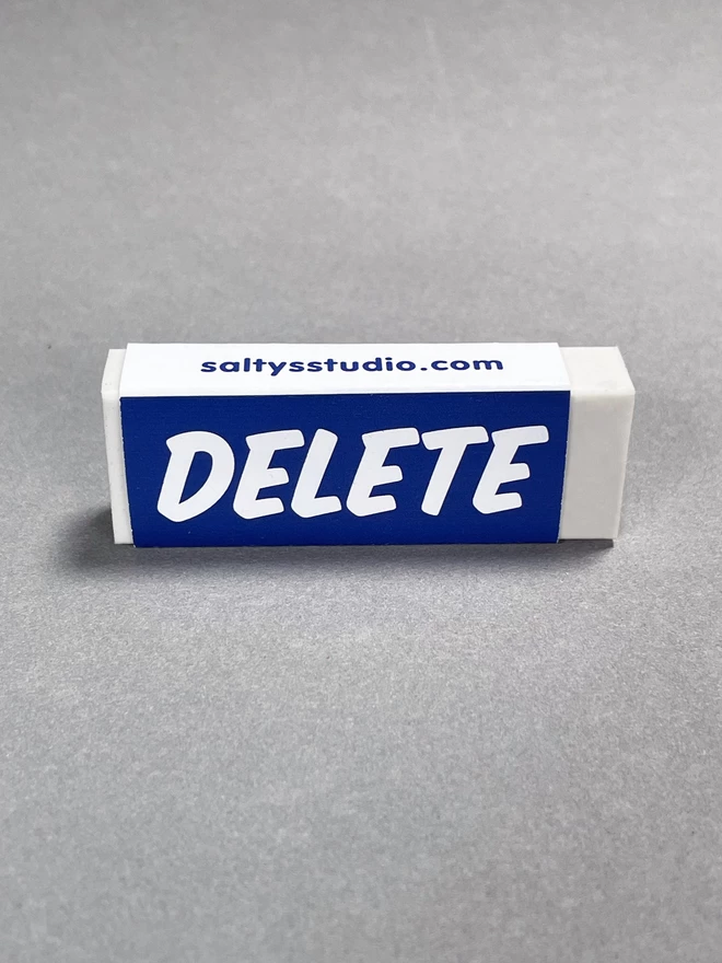 A delete rubber stands on its side. The rubber has a paper wrap around it with the words DELETE printed upon.