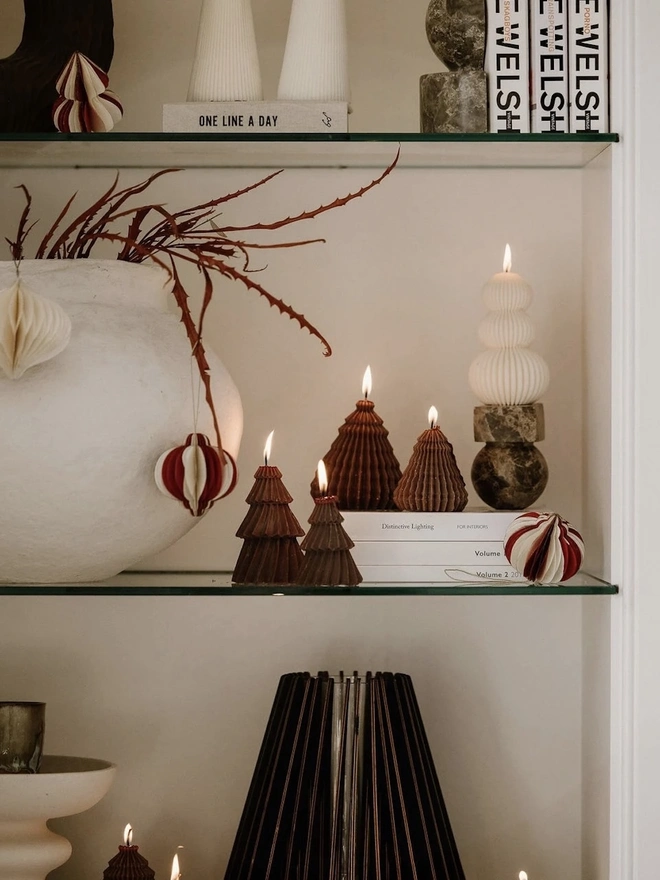 Brown Christmas Tree Warm Candles (Set Of 2). Glass shelves styled with lit chocolate brown Christmas tree candles, cream ribbed totem candles and swirled pillars, alongside a textured white vase, paper baubles and stacked books. The sculptural candles add warmth and a modern festive touch to a styled alcove.