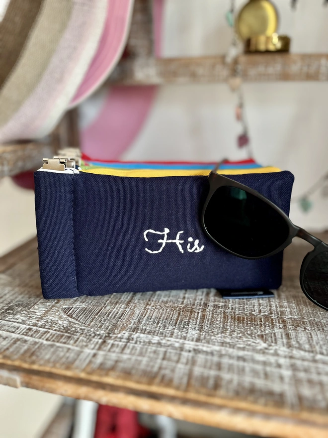Navy cotton spectacles case with 'His' embroidered on it. Sunglasses are draped over the pouch and a stack of pouches in different bright colours behind.