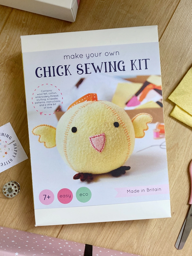 A craft kit to make a round yellow chick toy is on a wooden desk with some contents beside it.
