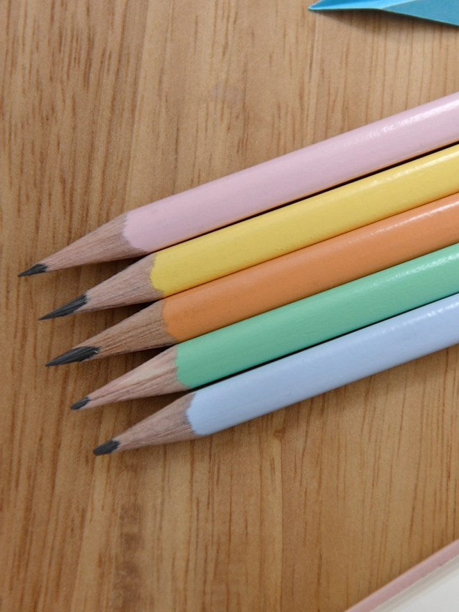 Five sharpened pastel coloured pencils lay on a wooden desk.