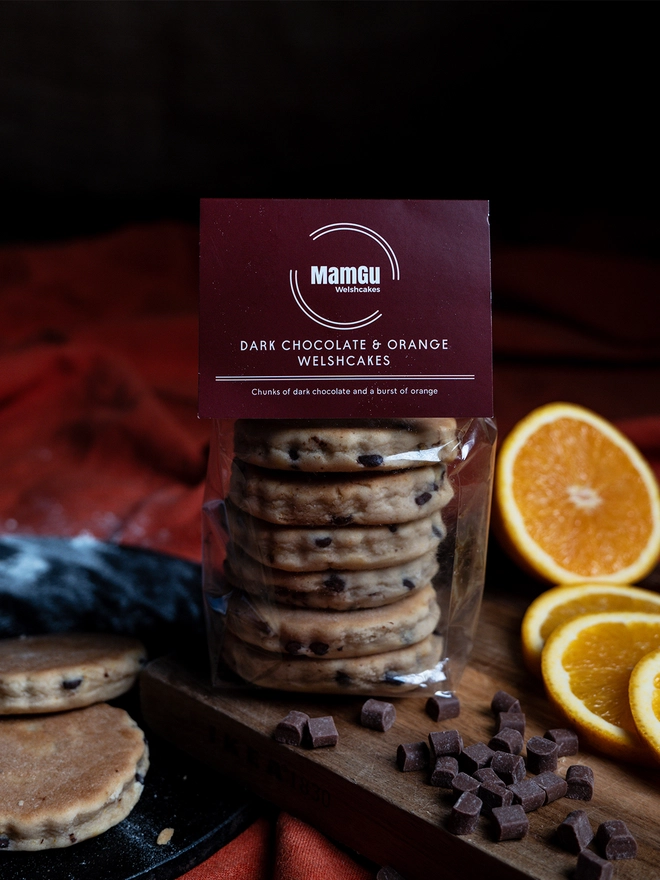 Packaged Dark Chocolate and Orange Welsh Cakes on a wooden chopping board with chocolate chips and slices of orange 