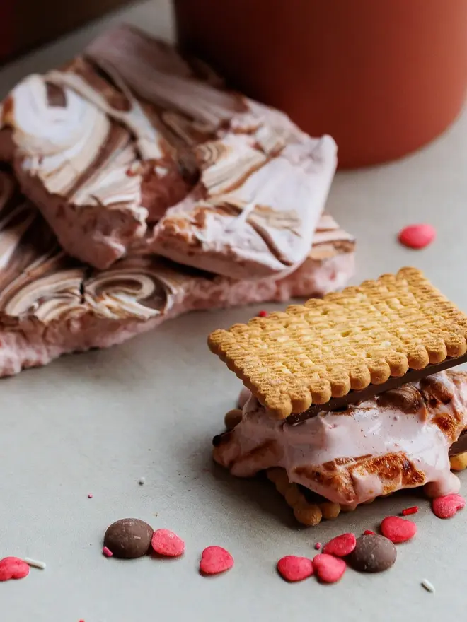 Date Night S'mores And Hot Chocolate Kit, a biscuit with wonder fluff sandwiched in the middle on a marble surface. It is near other sprinkles and items from the kit. 
