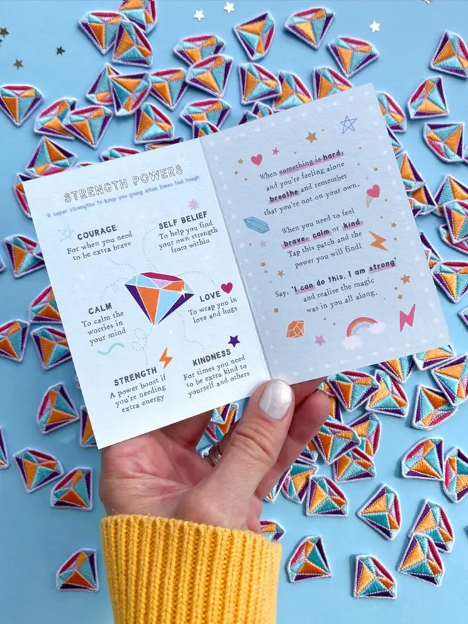 A hand holds open the Strength Powers card, showing the six powers and a short poem about feeling brave. Dozens of embroidered gem patches are scattered on a blue surface below.