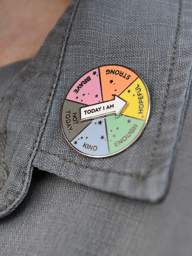 A round enamel pin that looks like a pie chart with six segments, each with positive word, and a white arrow that reads "Today I Am", is pinned to a grey shirt collar.