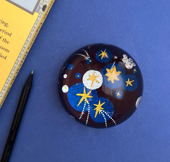 Glossy dome-shaped paperweight with gold stars on a dark blue background on a blue desk with a note pad in the background. 