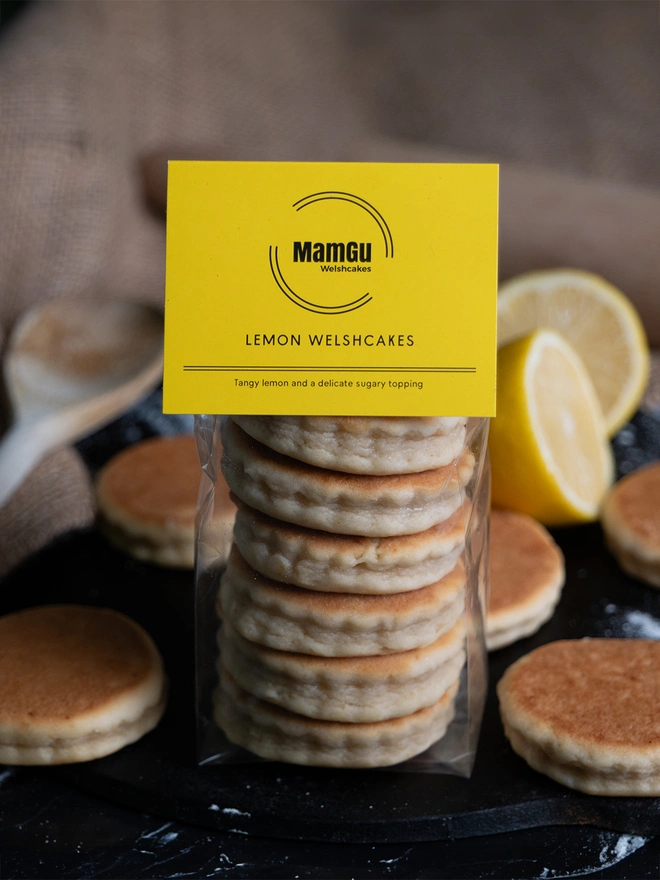 Packaged Lemon Welsh Cakes with a yellow MamGu Welshcakes label, sitting on a black griddle surrounded by loose welsh cakes and cut lemons