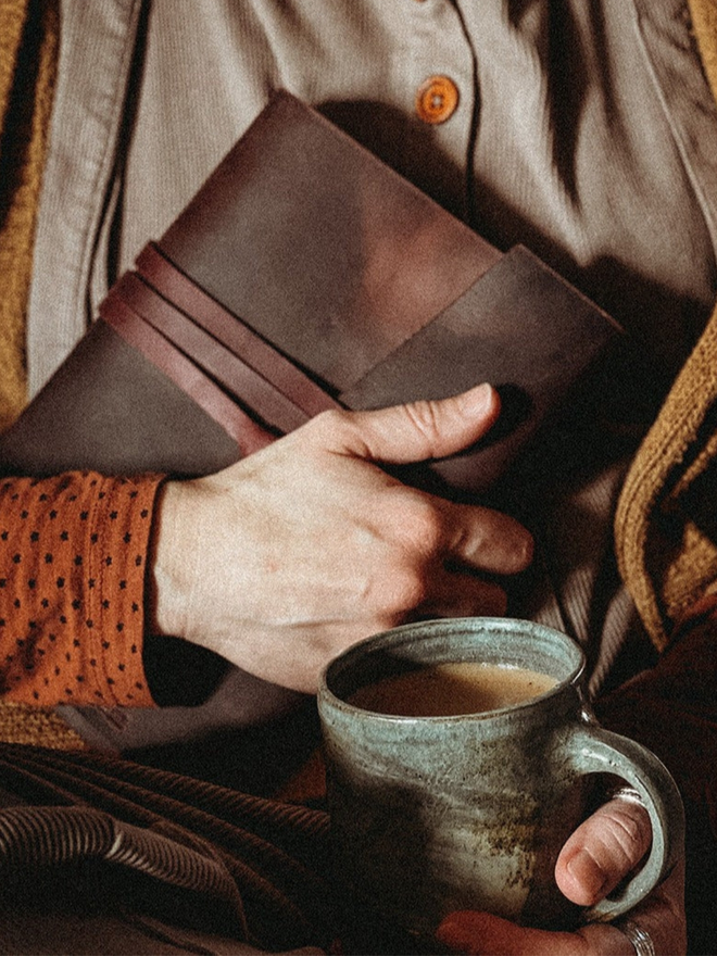 leather bullet journal. a dark brown leather journal in one hand, an cup of tea in the other.