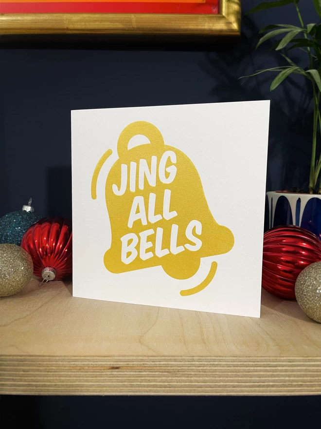 Sparkly gold ink graphic of a bell with the words Jing All Bells within. A square card on a plywood shelf with red and gold baubles around and a dark blue wall.