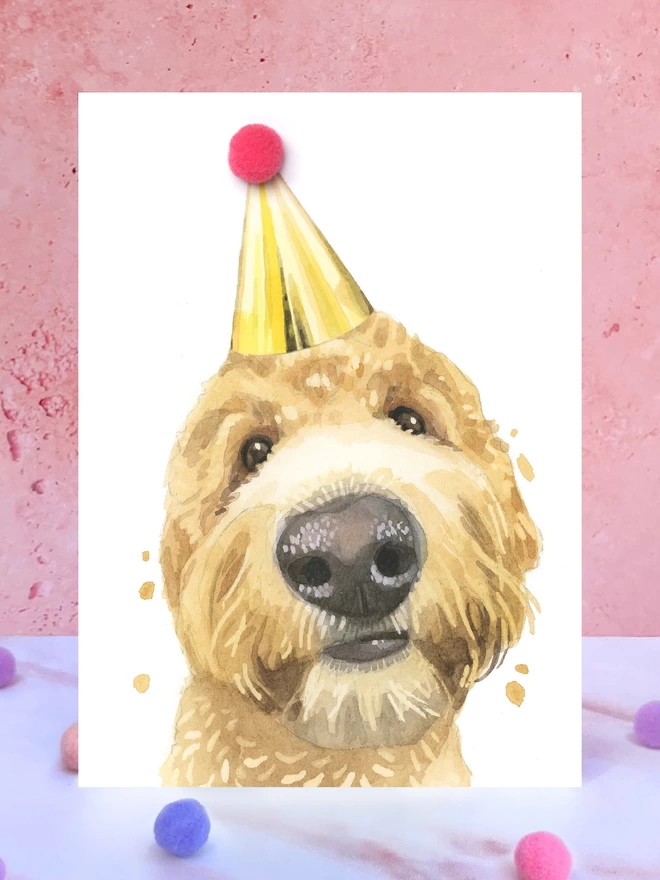 A greeting card featuring a hand painted design of a golden doodle, stood upright on a marble surface surrounded by pompoms. 