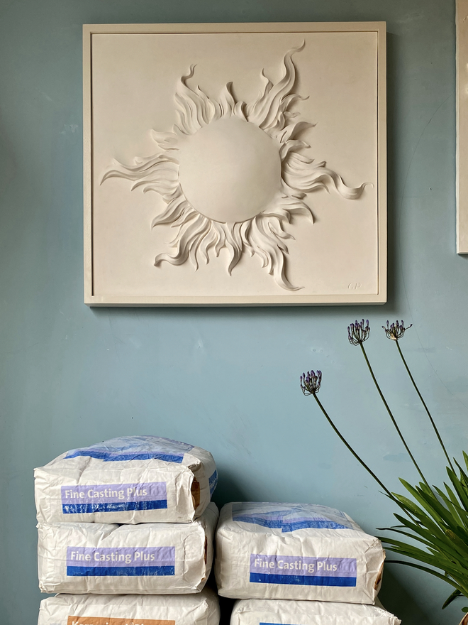 Bas-relief wall sculpture of a sun on a blue wall with bags of casting plaster and a houseplant