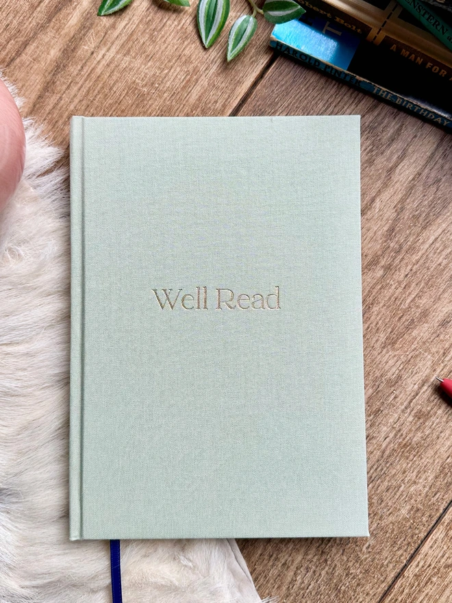 A green linen reading journal lies on a sheepskin rug. In the back ground are some really old books