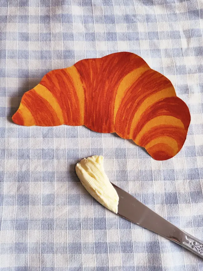  paper croissant painting on gingham table cloth, with butter knife and butter