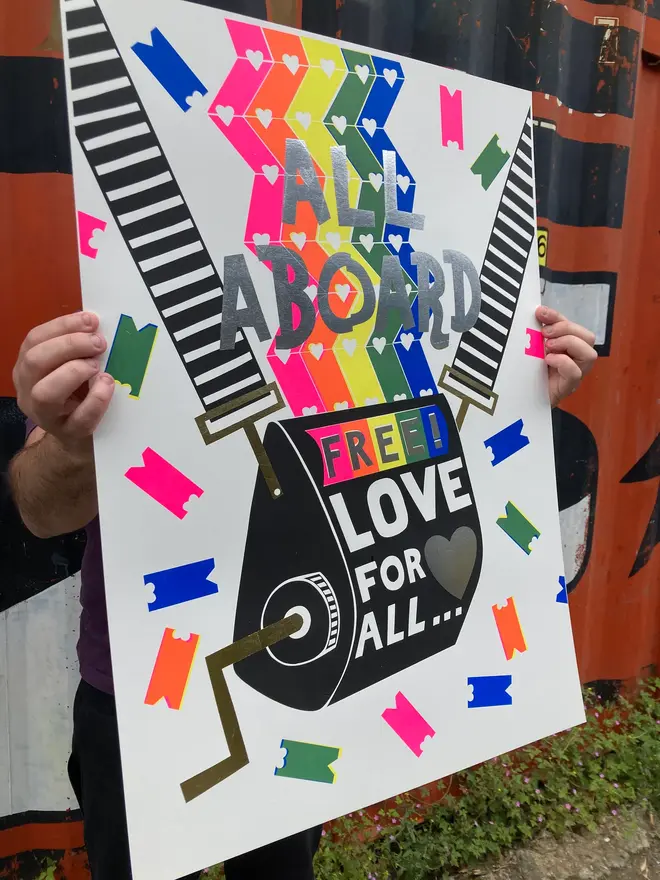 'All Aboard' Screenprint, a person holding a colourful screenprint in front of a decorated backdrop. The screenprint reads 'All aboard, free love for all'. 