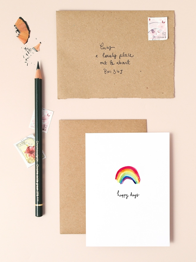 An image of a mini, rectangular white greetings card with a central illustration of a rainbow with a hand written 'happy days' message in black ink seen under the central illustration. The card is seen on top of a brown rectangular kraft envelope.