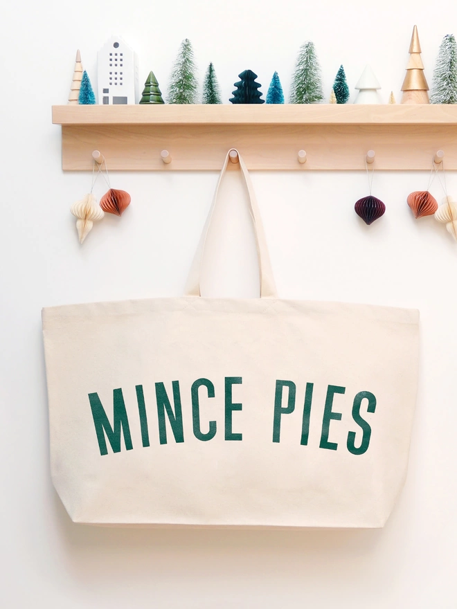 an oversized natural canvas tote bag with bold mince pies slogan hanging on a hook
