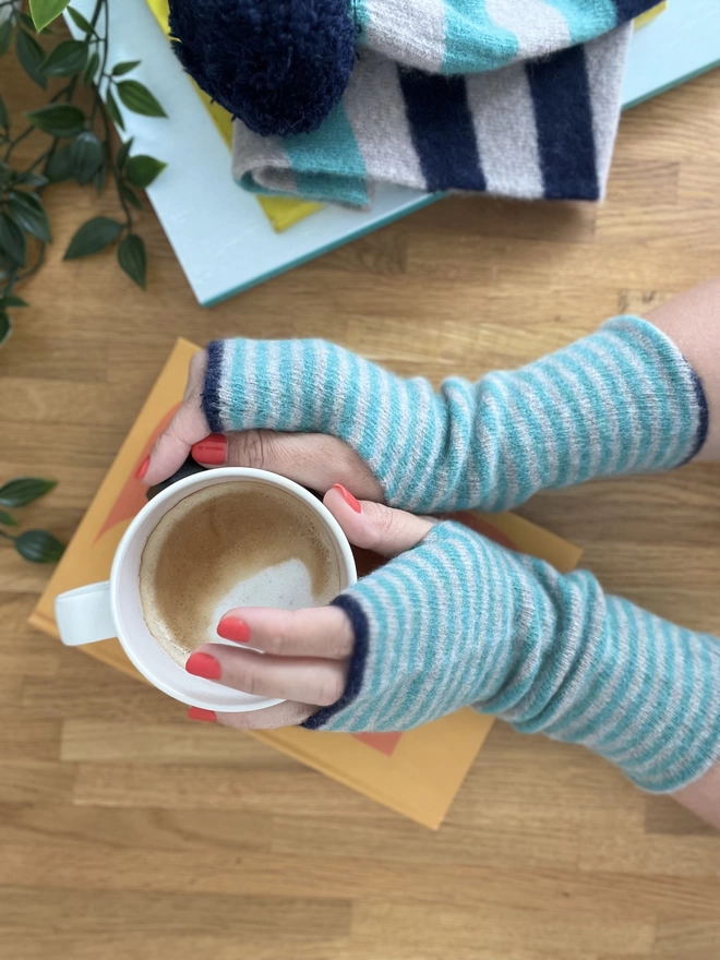 Knitted stripped wristwarmers being worn holding a coffee cup on onto of a book
