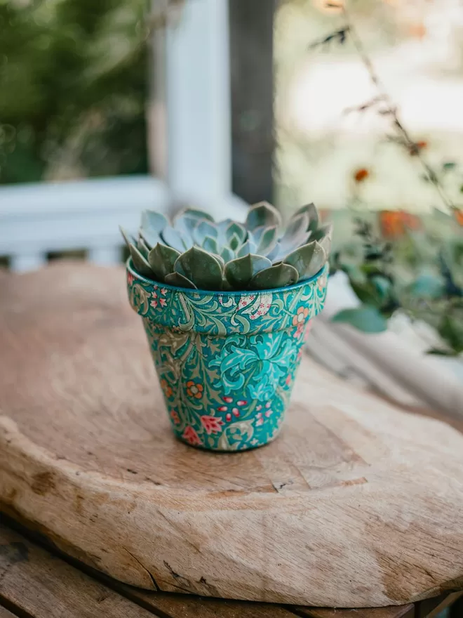William Morris Golden Lily Design Plant Pot Green And Pink seen on a wooden board outside.