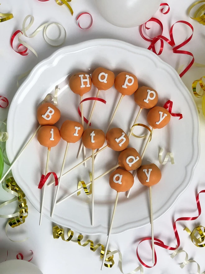 'Happy Birthday' Cake Pops, cake pops that say 'happy birthday' in white letters laid out onto a plate. It is surrounded by more birthday decorations.