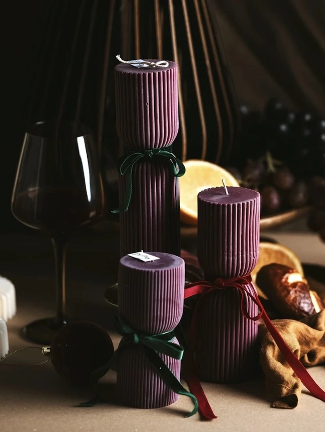 Burgundy Christmas Cracker Pillar Candles (Set Of 3). Ribbed plum pillar candles with cinched waists and velvet bows, styled beside a glass of red wine, orange halves and a velvet bauble on a moody table. The deep festive colour and sculptural shape bring warmth and elegance to winter entertaining.