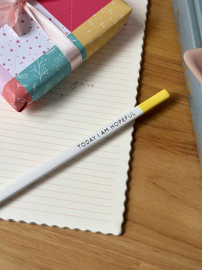 A white pencil with a yellow end and the words "Today I Am Hopeful" along the side lays on an open lined notebook, which rests on a wooden desk.