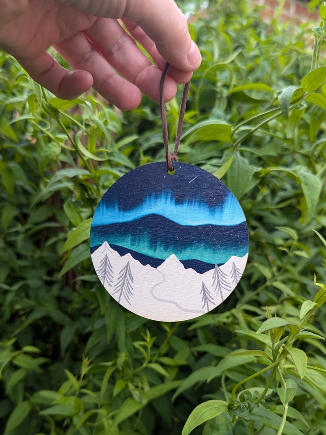 A wooden circle decoration with the northern lights over a white snowy mountain. Measures roughly 10 inches by 10 inches, with a suede vegan hoop for hanging. Illustrated by Charis Raine.