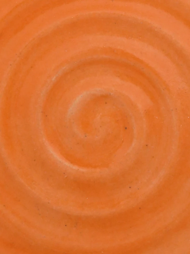 A close-up of a handmade ceramic glaze swatch in mandarin orange, showing a smooth concentric swirl texture with a delicate, even tone throughout