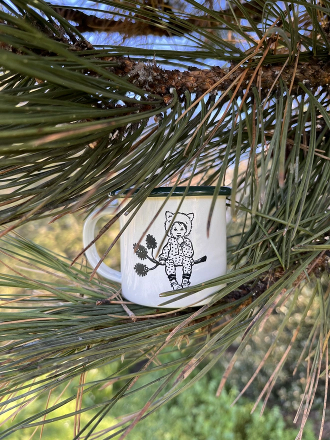a miniature enamel mug sat in a pine tree the mug is made from enamel it is cream with a green rim and features an etched illustration of a small pixie child wearing a wolf hood