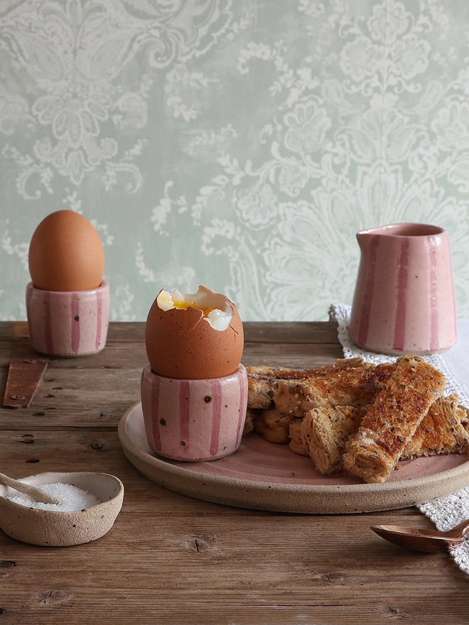 Two pink-striped ceramic egg cups holding boiled eggs sit on a wooden table alongside a matching plate of buttered toast and a small milk jug.
