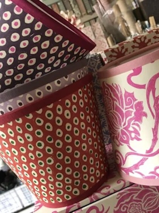 Candle Clip Shades. A collection of small hand block printed lampshades in plum spot, red spot and pink floral designs displayed in a paper studio. The colourful mix of patterns shows the range of shades available for wall lights and table lamps.
