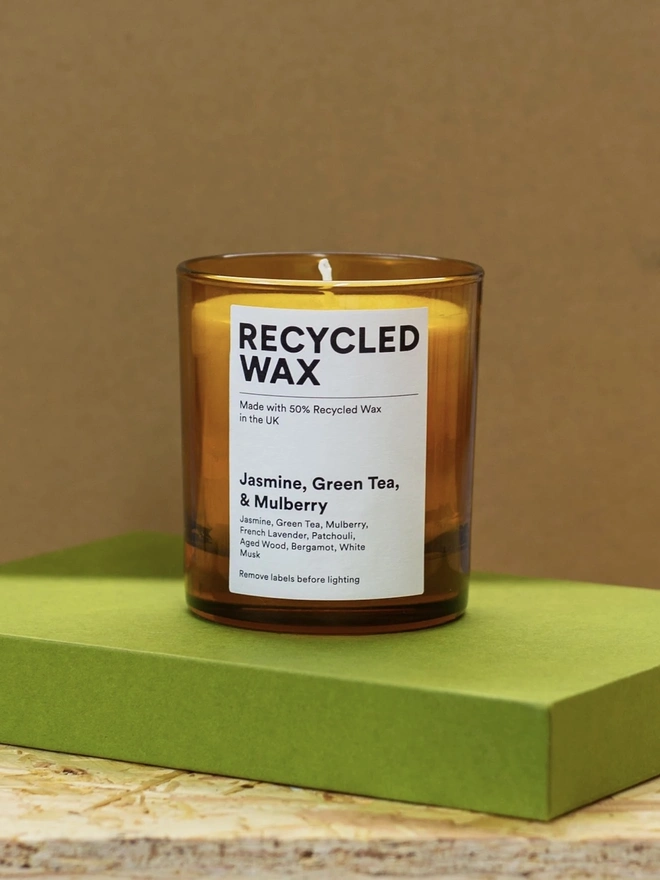 Glass Scented Candle. the jasmine, green tea and mulberry glass scented candle stands on a green box against a brown background, housed in an amber glass jar with a single wick. a white label reads "recycled wax, made with 50% recycled wax in the uk", followed by the scent name and note list of jasmine, green tea, mulberry, french lavender, patchouli, aged wood, bergamot and white musk. the warm amber glass gives the candle a simple, understated finish.
