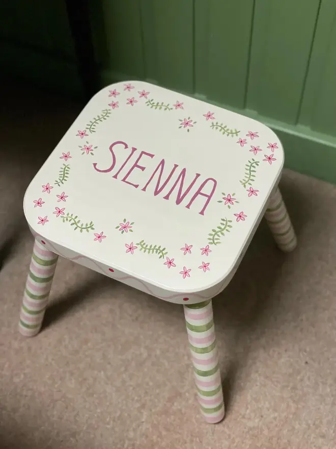 personalised hand-painted nursery stool. a small white stool with green and pink striped legs, painted with 'sienna' on top.
