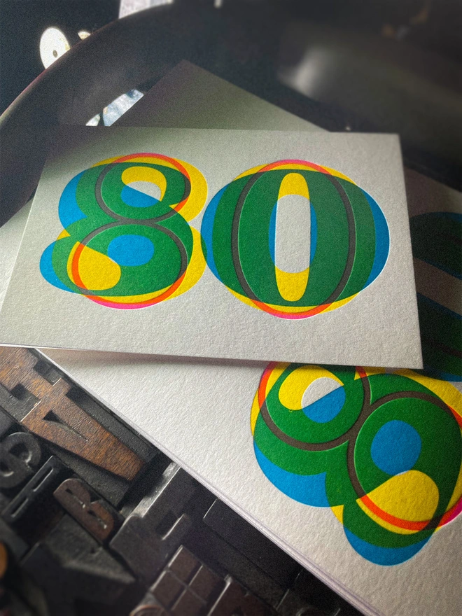 Handmade letterpress 80th birthday card featuring bold overlapping numerals printed in vibrant fluorescent cyan, magenta and yellow inks. Each card is individually hand printed using traditional letterpress techniques on luxury paper stock, creating a beautifully tactile finish with a subtle deboss and rich layered colours inspired by the CMYK printing process.