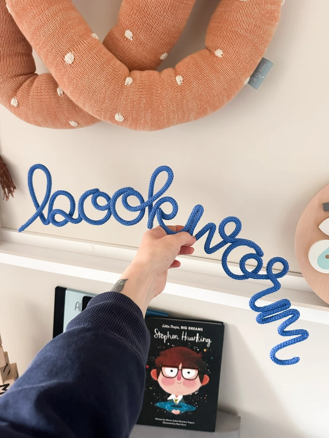 bookworm knitted wire word sign being held up infront of a white shelf in a kids playroom or bedroom area with some childrens books in a reading area.