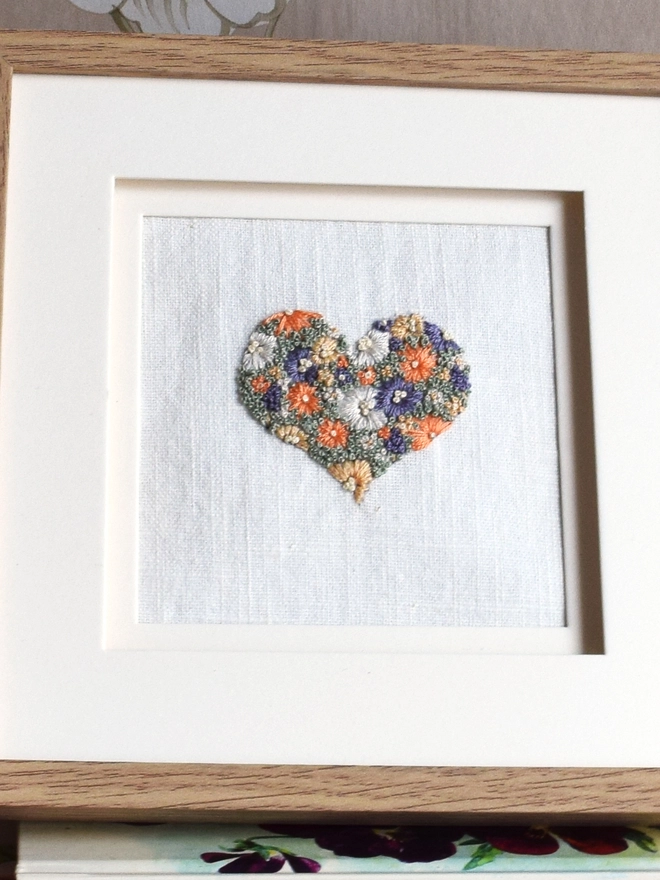 An embroidered Sunshine Garden Heart, of Golden Yellows and Bright Orange Blossoms with Green French Knot grass background.  Displayed in an oak frame