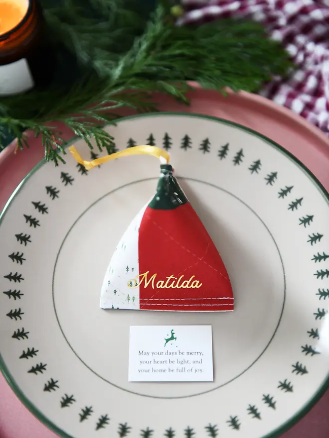 A traditional patchwork design fabric fortune cookie with gold personalised name “Matilda” sits on a Christmas dinner plate, ready as a handmade festive place setting with a hidden Christmas wish card beneath.