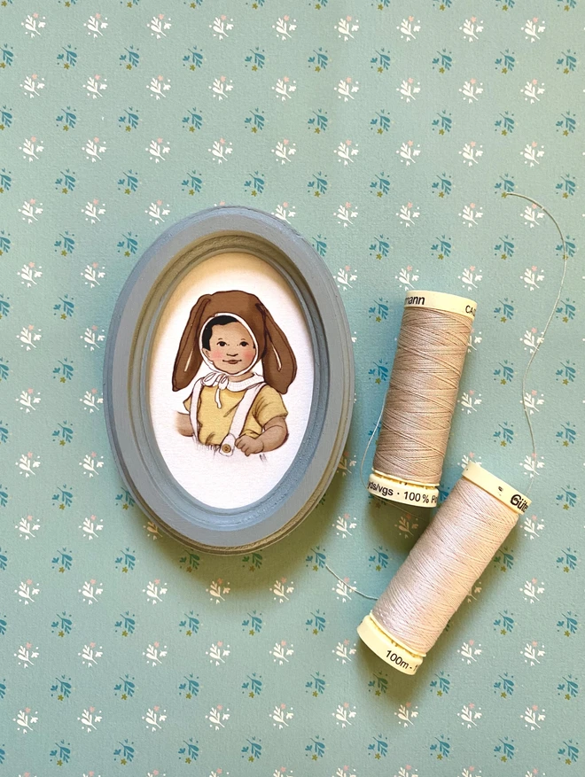 a photograph of a tiny miniature farmed personalised portrait of an asian boy waering a easter bonnet laid next to 2 sewing threads