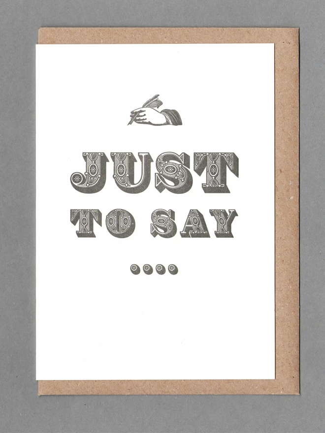 White card with a black hand and black text reading 'JUST TO SAY...' with a kraft envelope behind it
