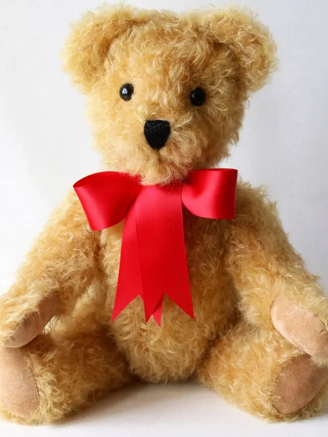 Handmade brown teddy bear sitting upright with a red ribbon bow against a plain background.