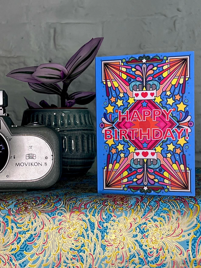 A vibrant blue birthday card with a bold multi-coloured pattern, with Happy Birthday at the centre, sits on a shelf covered in a golf and blue wrapping paper, next to a purple houseplant in a green pot, and a vintage silver camera.