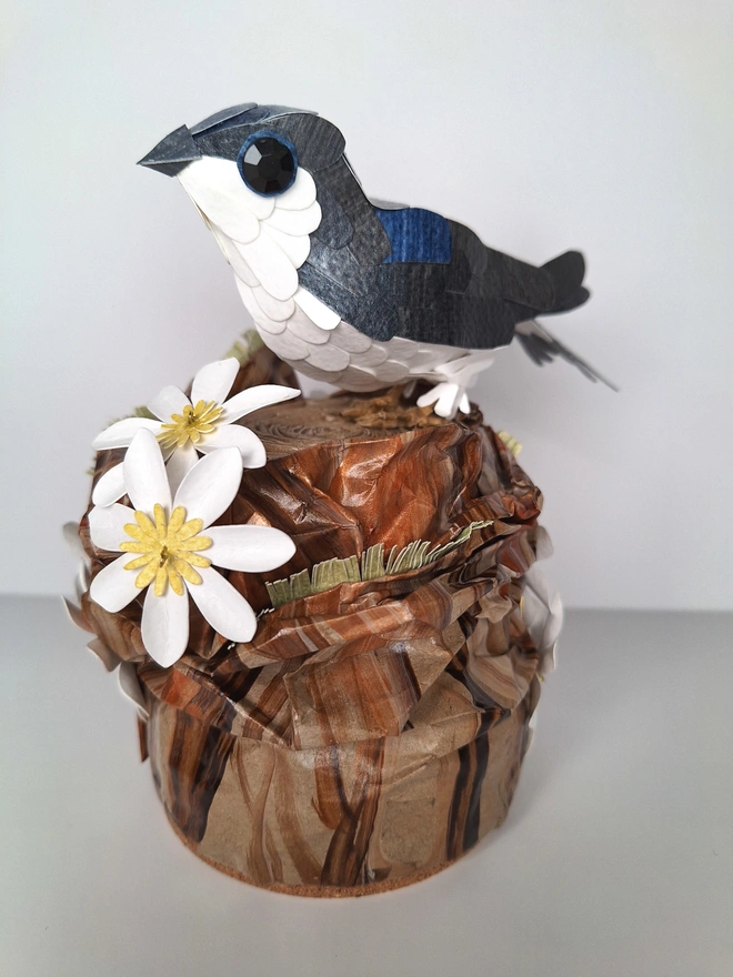 Housemartin bird sculpture, perched on a floral tree stump. This handmade sculpture is made from recycled paper. The bird is black and blue, with a white belly. White and yellow flowers adorn the tree stump.  Side view.